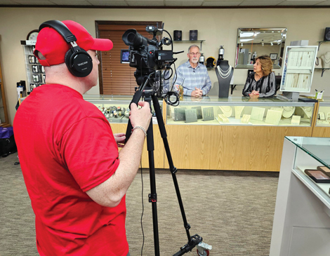 A man in red filming a couple standing behind a jewelry counter