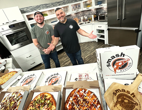 Two men smiling and pointing to a spread of pizza in boxes from Smash Pizza Kitchen in North Ridgeville, OH